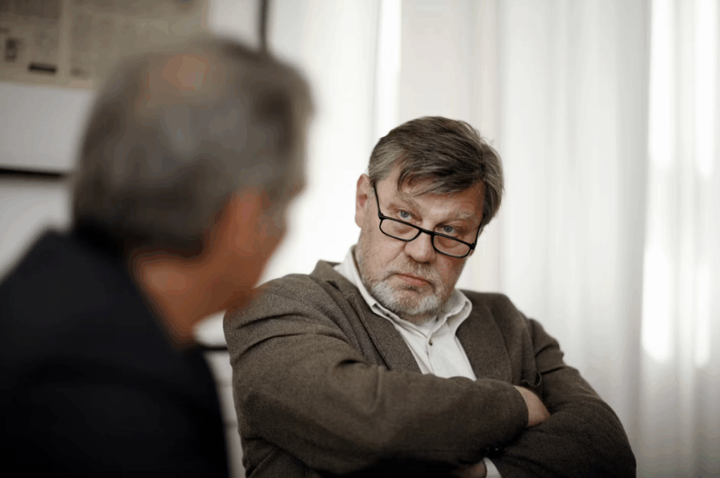Two men sit and talk. One wears a brown sweater and glasses. The other a black coat with his back to the camera.