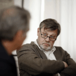Two men sit and talk. One wears a brown sweater and glasses. The other a black coat with his back to the camera.