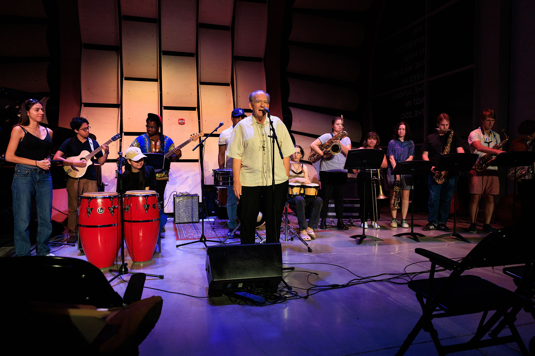A large audience in a dark auditorium smiles and waves at the camera, with a bright light shining from the back of the room. The crowd appears cheerful and diverse, filling nearly every seat. There a warm, welcoming atmosphere.
