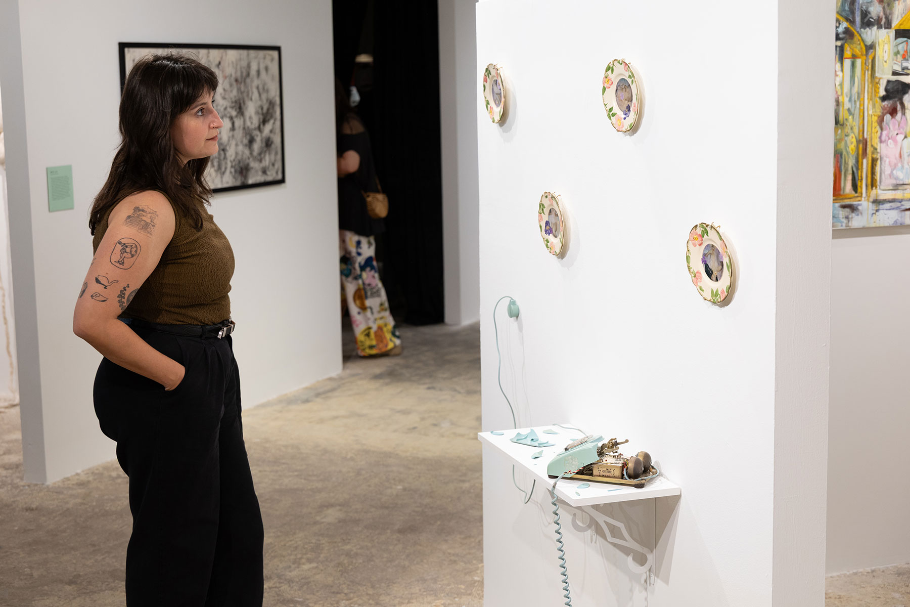 A woman with tattoos on her arm stands in an art gallery, observing a wall-mounted installation of five decorative plates, each featuring a portrait surrounded by floral designs. Below the plates is a white shelf holding a vintage rotary telephone and various small objects. Other artworks and people are visible in the background.