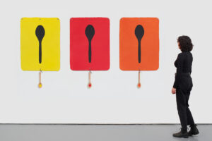 A woman in black looks at three artworks of spoons with spoons hanging from the bottom of the artworks.