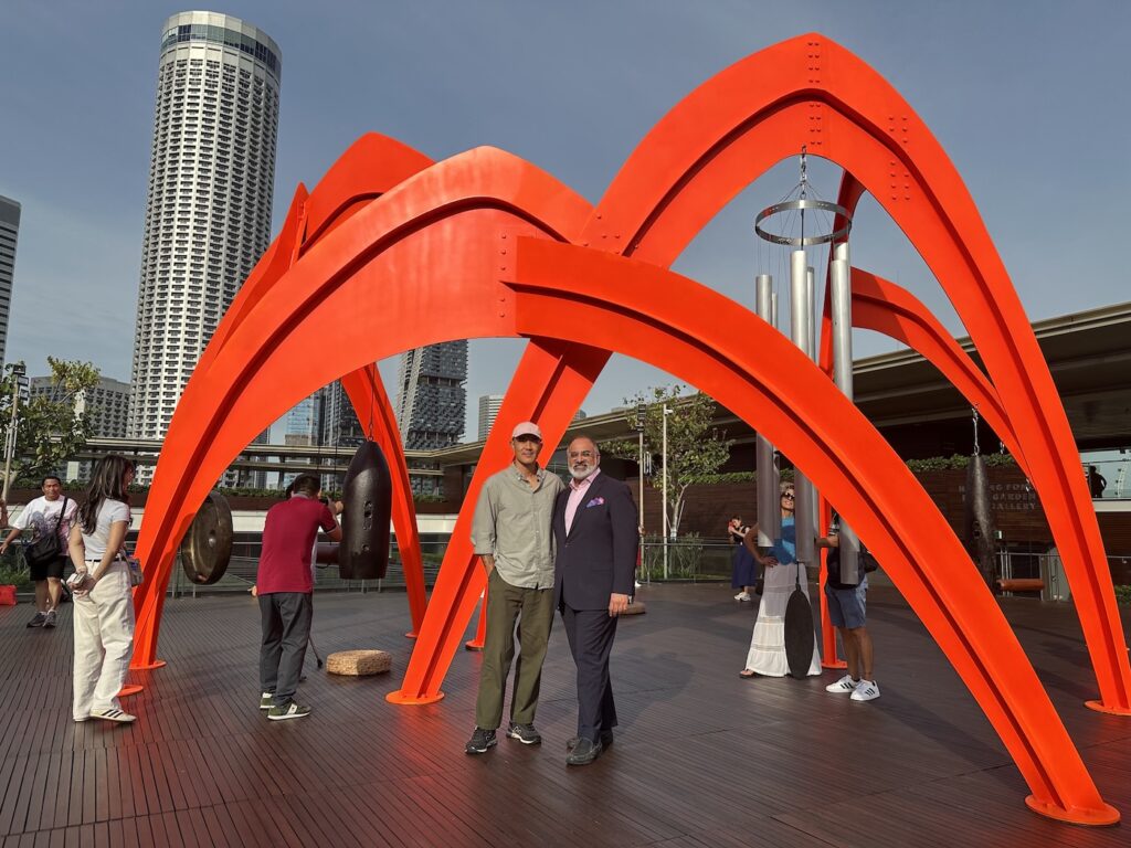 Two men posing under a massive, bright red arched metal sculpture, resembling a spider, situated on an outdoor wooden deck in front of a modern skyscraper and other city buildings. The sculpture has large gongs and wind chimes hanging from its frame.