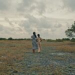 Two women in dresses walk away from the camera across a dry, grassy field toward a large green tree on the right under a cloudy, overcast sky.