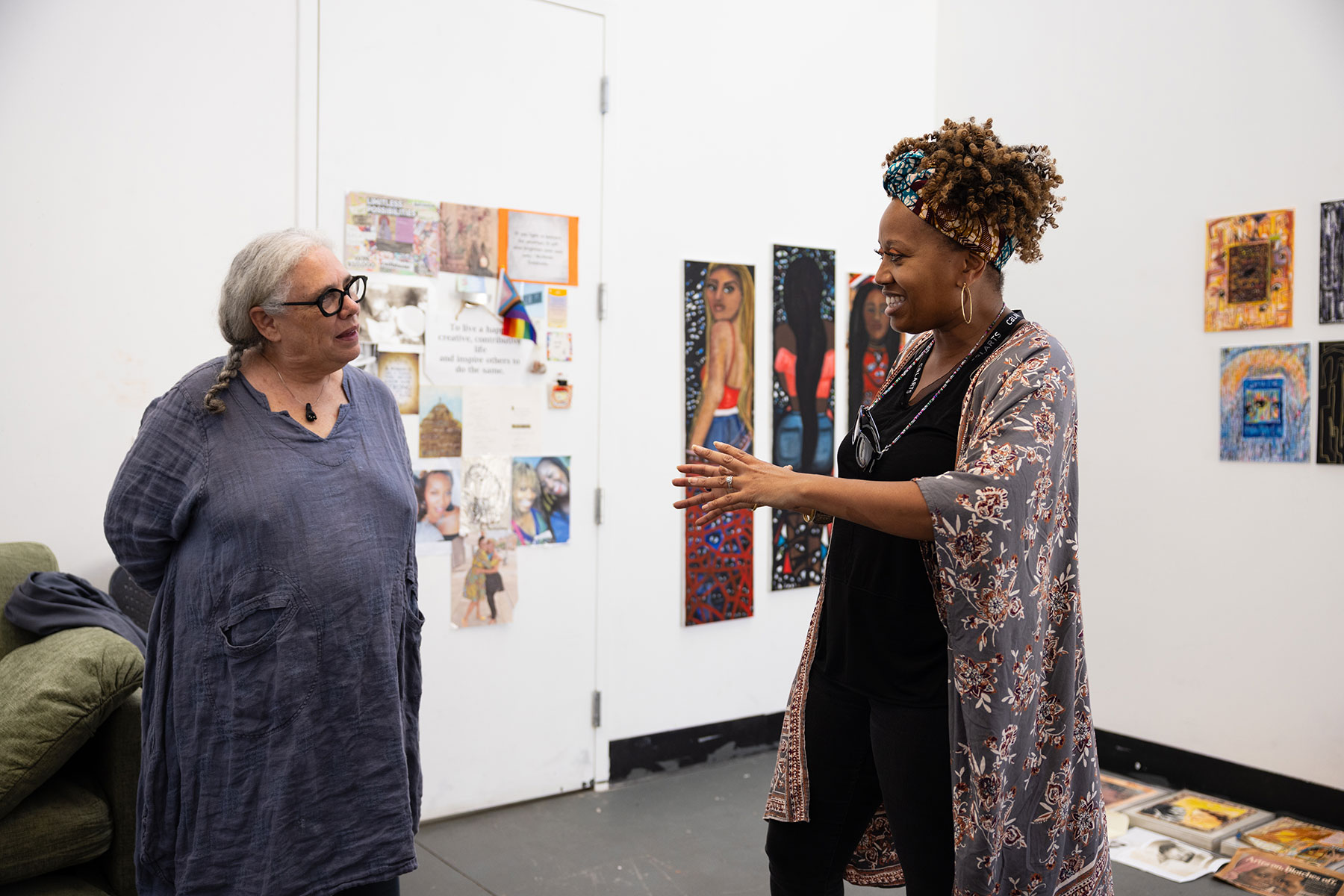 Visiting artist Alison Saar and a student engage in an animated conversation inside a bright studio space. The walls are adorned with vibrant paintings and mixed-media pieces, while art supplies and works-in-progress line the floor. The moment captures mentorship, artistic dialogue, and the collaborative learning environment central to CalArts.