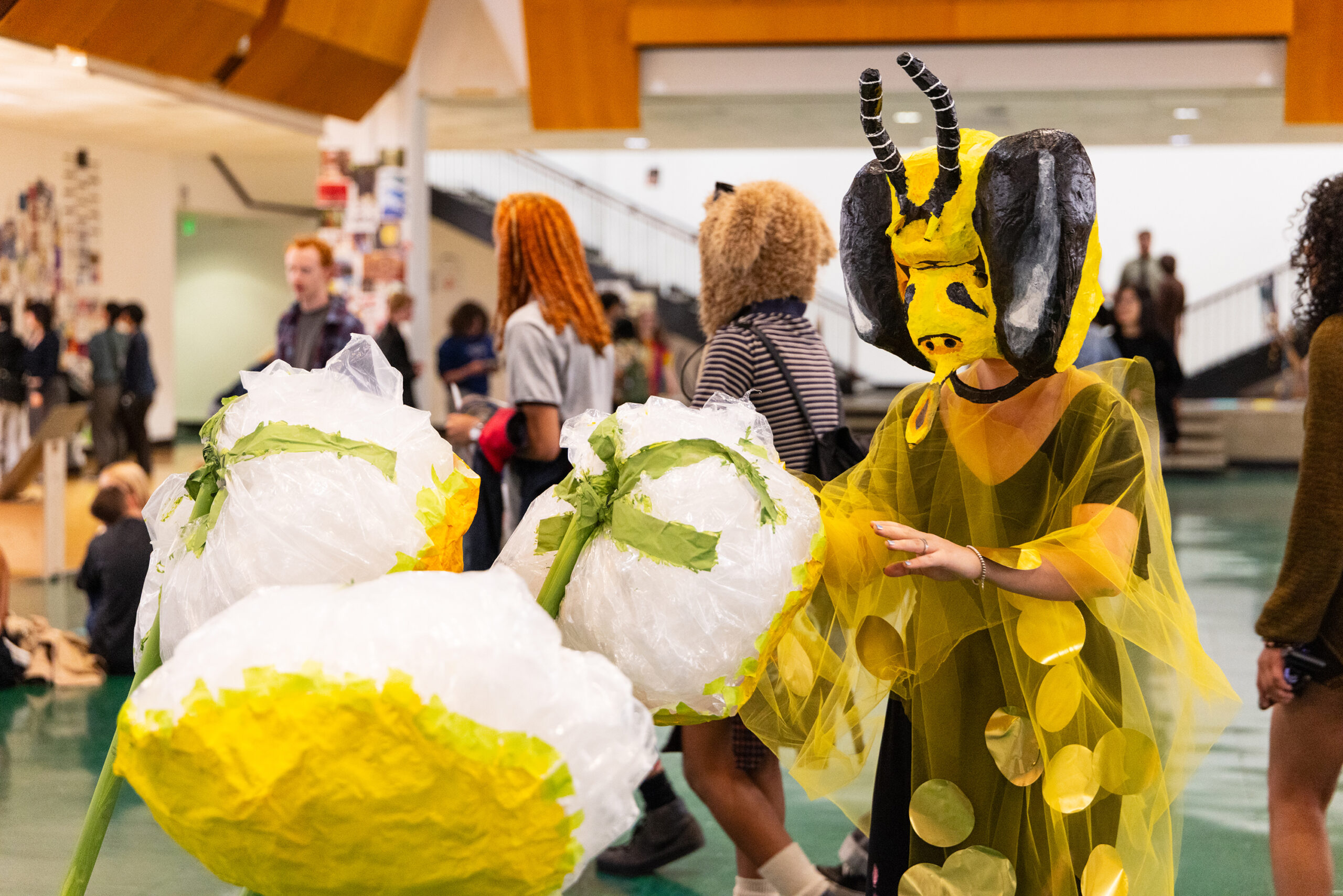 A CalArts student wearing a handmade bee-inspired costume with a large yellow and black headpiece and translucent yellow fabric interacts with oversized white and yellow flower sculptures in the Institute’s main gallery area. Other students and visitors are seen in the background, adding to the vibrant, communal energy of the space. The image captures creativity, performance art, and student engagement at CalArts.