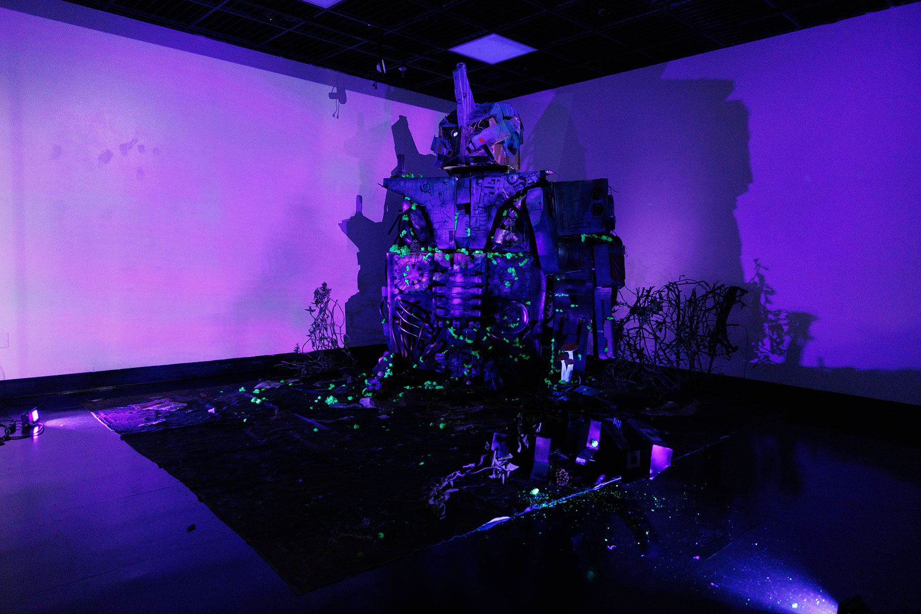 A dark room illuminated by purple and blacklight reveals a large sculptural installation resembling a robot or mechanical figure made from repurposed materials. Neon green elements glow on the sculpture and floor, and twisted wires and debris surround the piece, creating an eerie, futuristic atmosphere.