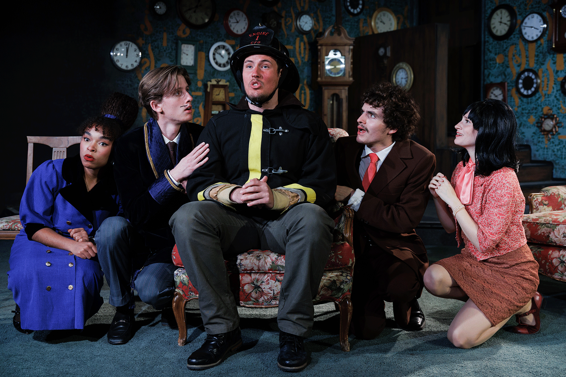 A group of five actors in colorful, vintage-inspired costumes gather closely around a seated firefighter character onstage. The background is filled with clocks of various sizes. The actors gesture expressively toward the firefighter, creating a humorous and dramatic tableau.