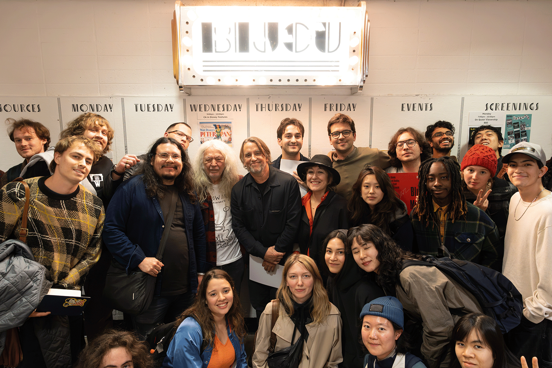 A large group of students and faculty stand closely together smiling in front of a bright “BIJOU” marquee sign. The group appears energetic and joyful, posing for a photo in a hallway with posters and schedules on the wall behind them.