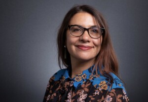 A woman with long, dark brown hair and glasses smiles against a dark gray background. She is wearing a brown floral blouse with a blue patterned collar and shoulder detail.