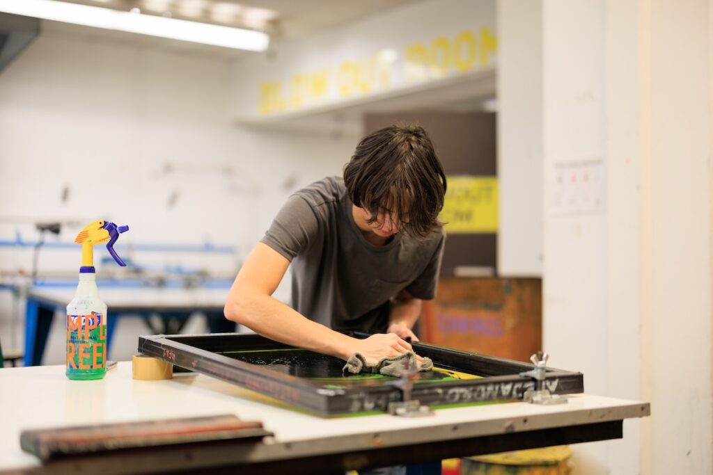 A young person cleans a screen-printing frame with a rag on a white studio table, with a spray bottle of green cleaner in the foreground.
