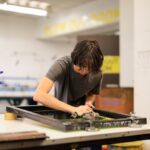 A young person cleans a screen-printing frame with a rag on a white studio table, with a spray bottle of green cleaner in the foreground.