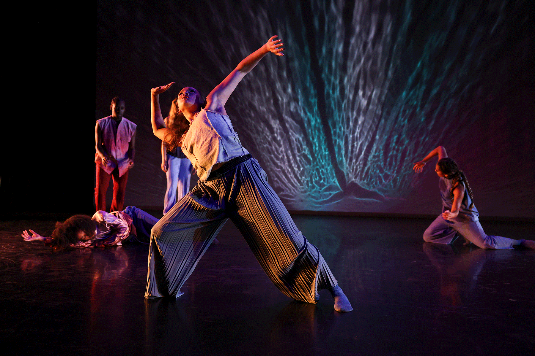 Several dancers perform on a dimly lit stage with vibrant, multicolored lighting projected behind them in a pattern resembling flowing water. A dancer in wide pleated pants reaches upward in a dramatic pose while others move or lie on the floor around her.