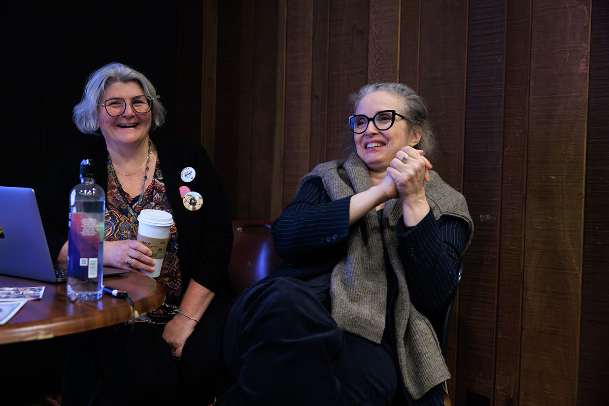 Two older women sit at a small round table in a wood-paneled room, smiling and engaged in conversation. One woman with short gray hair and glasses holds a takeaway coffee cup beside an open laptop and a water bottle, while the other woman, also wearing glasses and a layered outfit, sits with her hands clasped, appearing animated and amused.