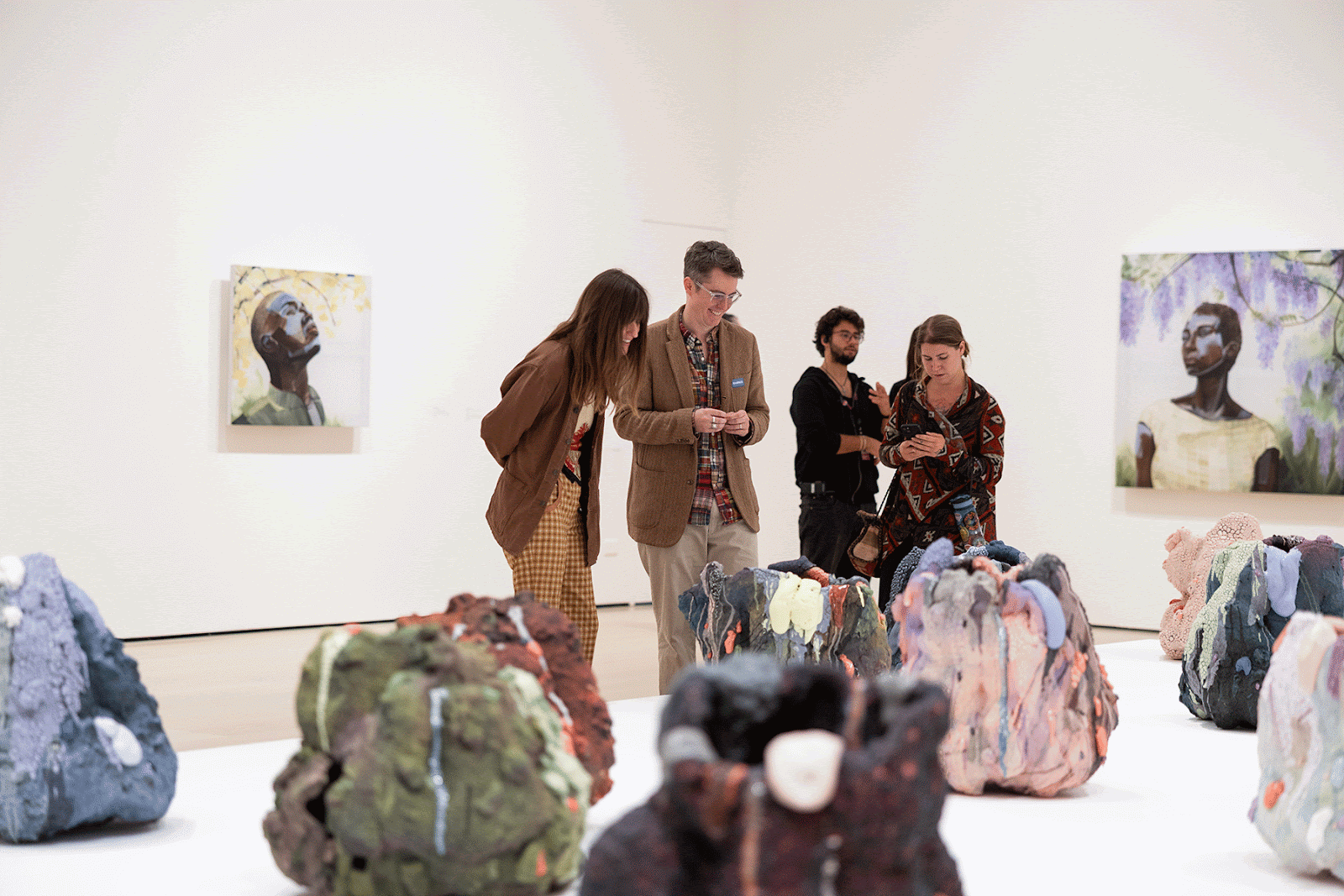 Visitors stand in a white-walled art gallery, closely examining a table of colorful, rock-like sculptural forms in the foreground. Two people lean in to discuss the artworks, while others nearby look at the pieces or check a phone. Framed portrait paintings hang on the walls behind them.