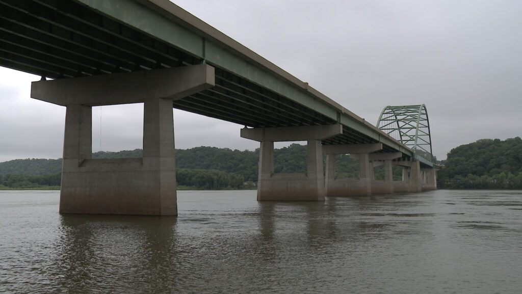 A bridge with green framework and concrete supports spans a wide river, with lush green hills in the background under a cloudy sky.