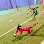 Miniature soccer scene from Fútbol Is Life featuring a goalkeeper in a red uniform diving to block a shot near the goal, with other players visible in the background on a detailed green pitch.