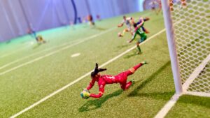 Miniature soccer scene from Fútbol Is Life featuring a goalkeeper in a red uniform diving to block a shot near the goal, with other players visible in the background on a detailed green pitch.