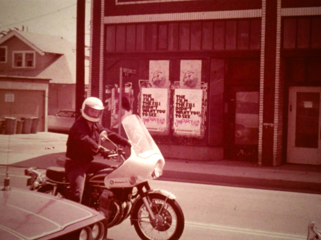 A person on a motorcycle in front of a building with posters in the windows.