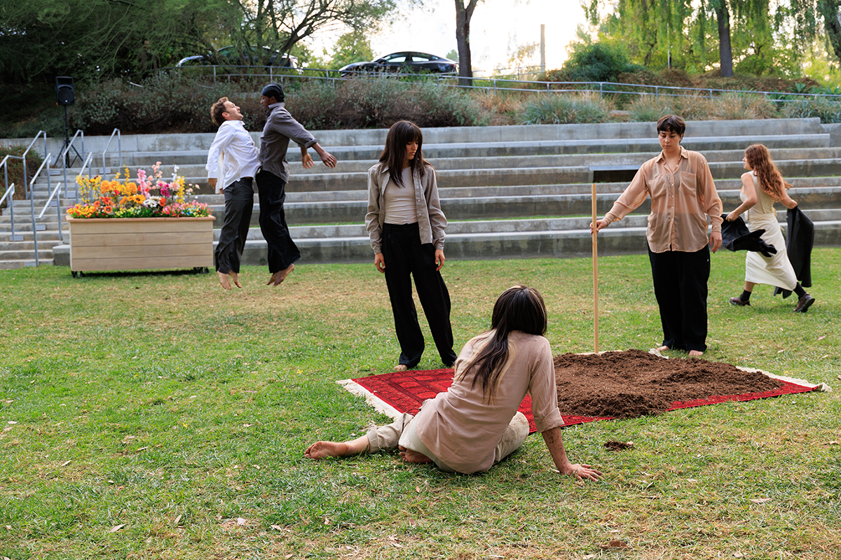 An outdoor performance shows several people arranged around a patch of soil on a grassy lawn, with some standing, one seated on the ground, and two jumping in midair near tiered concrete seating.