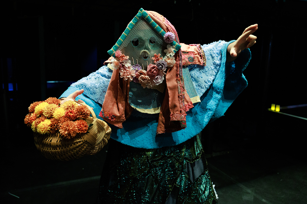A costumed performer stands on a dark stage wearing a blue, layered robe and a mesh face covering decorated with flowers. They hold a woven basket filled with orange and yellow flowers in one hand while extending the other arm outward, as if mid-gesture or dance.