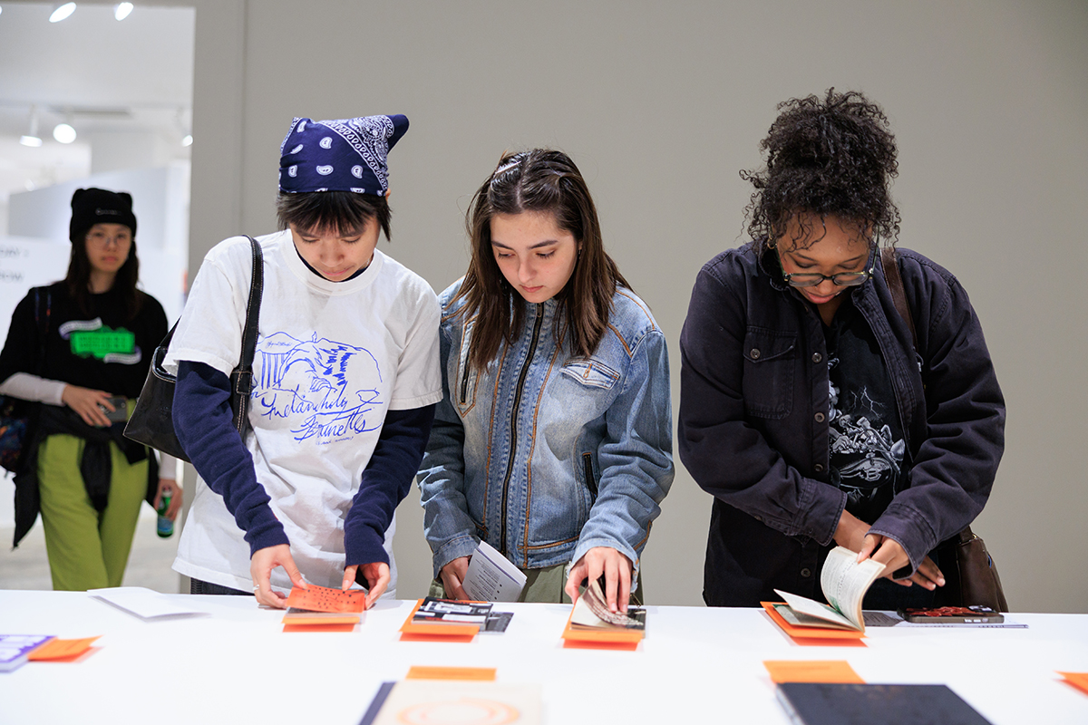 Four people stand at a long white table in a gallery, looking down and flipping through small printed booklets or pamphlets displayed in neat stacks.