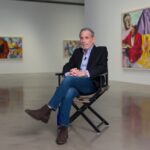 A man sits on a director's chair in the middle of a gallery.