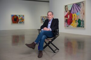 A man sits on a director's chair in the middle of a gallery.