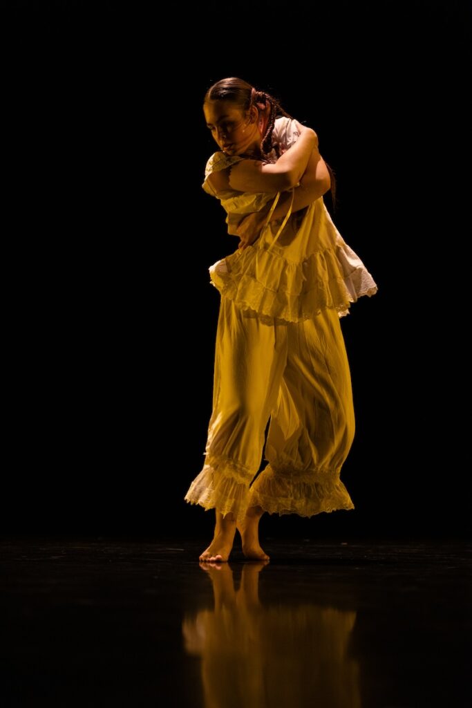 A woman in a mustard ensemble dances in a black studio.