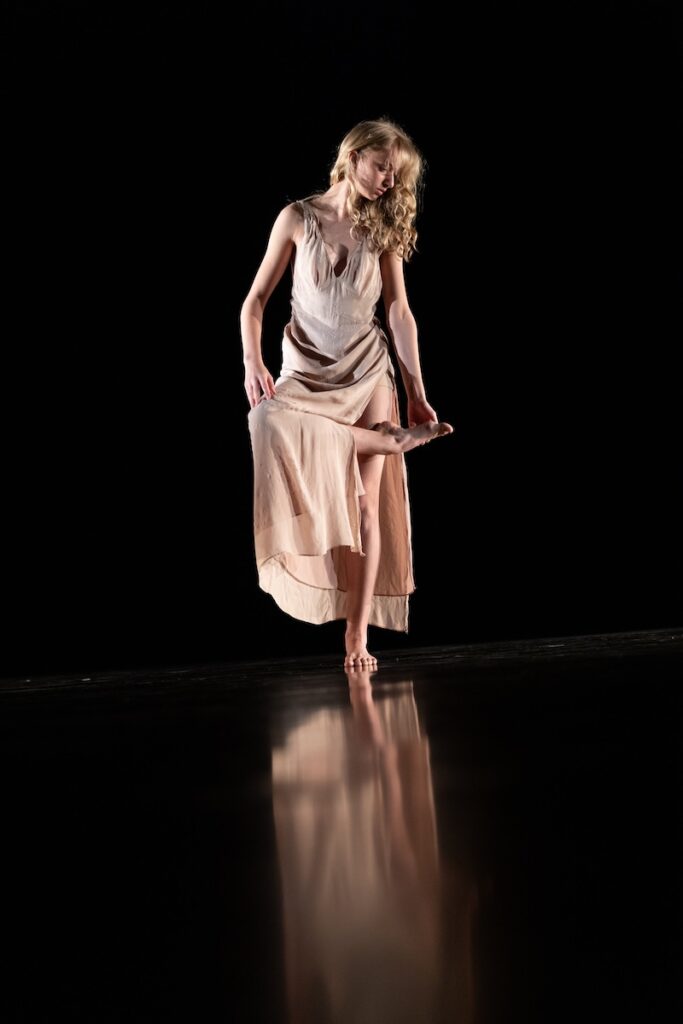 A woman in a cream dress dances on a black stage.