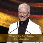 A screenshot of a smiling man at a podium. He is wearing a black turtleneck, a cream blazer and glasses. The overlay reads, "Winsor McCay Award / Chris Sanders."