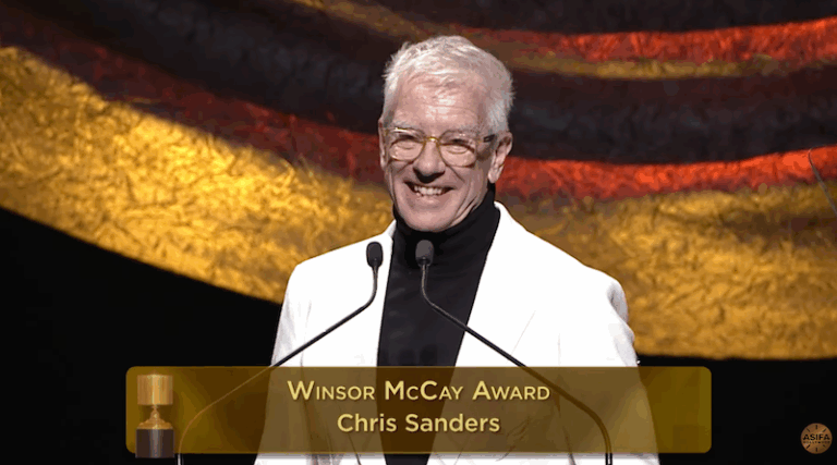 A screenshot of a smiling man at a podium. He is wearing a black turtleneck, a cream blazer and glasses. The overlay reads, "Winsor McCay Award / Chris Sanders."
