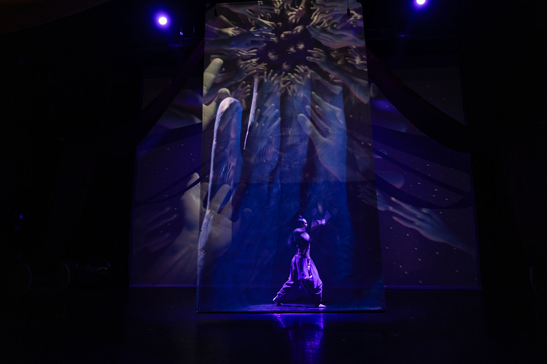 A solitary performer in purple clothing stands illuminated center stage against a projection displaying numerous reaching hands in dark blue and purple tones.