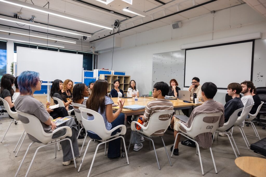 A group of people sitting around a table in a modern classroom, engaged in discussion.