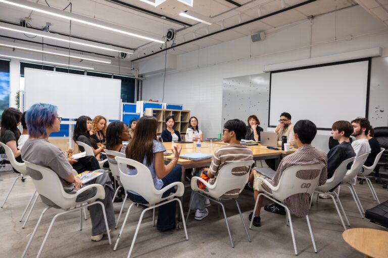 A group of people sitting around a table in a modern classroom, engaged in discussion.