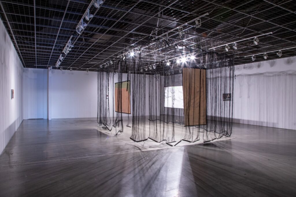 Gallery installation with hanging black sheer fabrics and rectangular painted canvases.