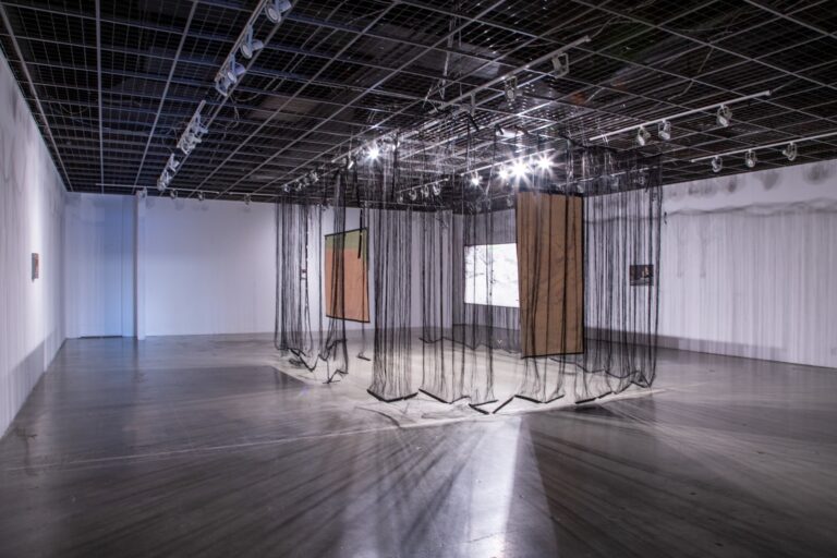 Gallery installation with hanging black sheer fabrics and rectangular painted canvases.