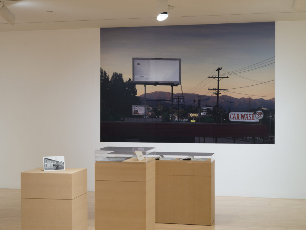 Gallery setting with a large photograph of a billboard and skyline at dusk, alongside two display pedestals. Transcribed Text: CAR WASH COIN OP Ralphs