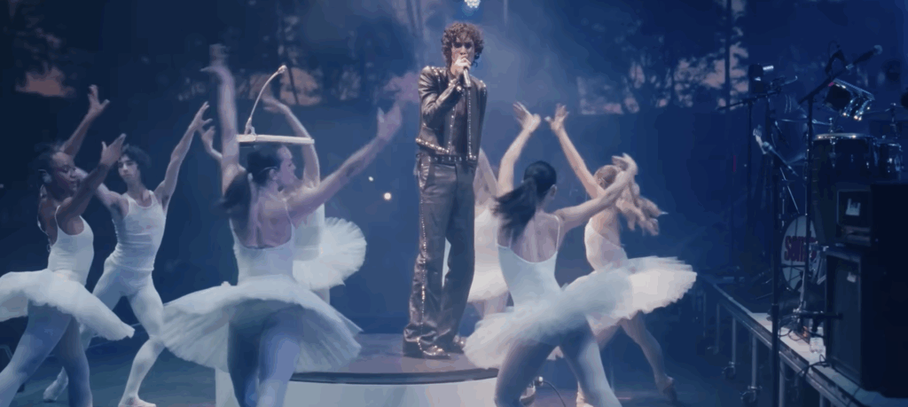 A singer in a studded leather outfit performs on stage surrounded by ballet dancers.