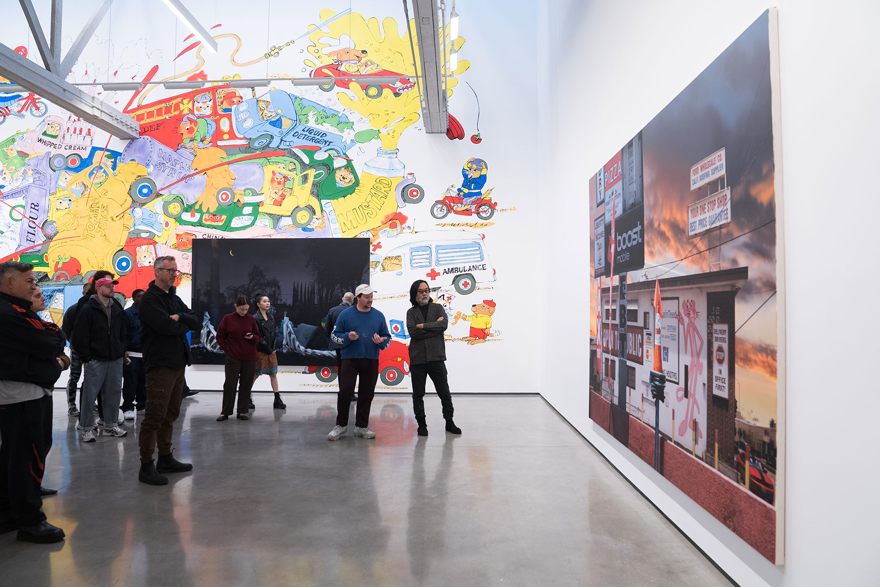 A group of people views contemporary art installations, including a large, colorful mural and a photograph of a storefront against a sunset sky, in a brightly lit gallery with a polished concrete floor.