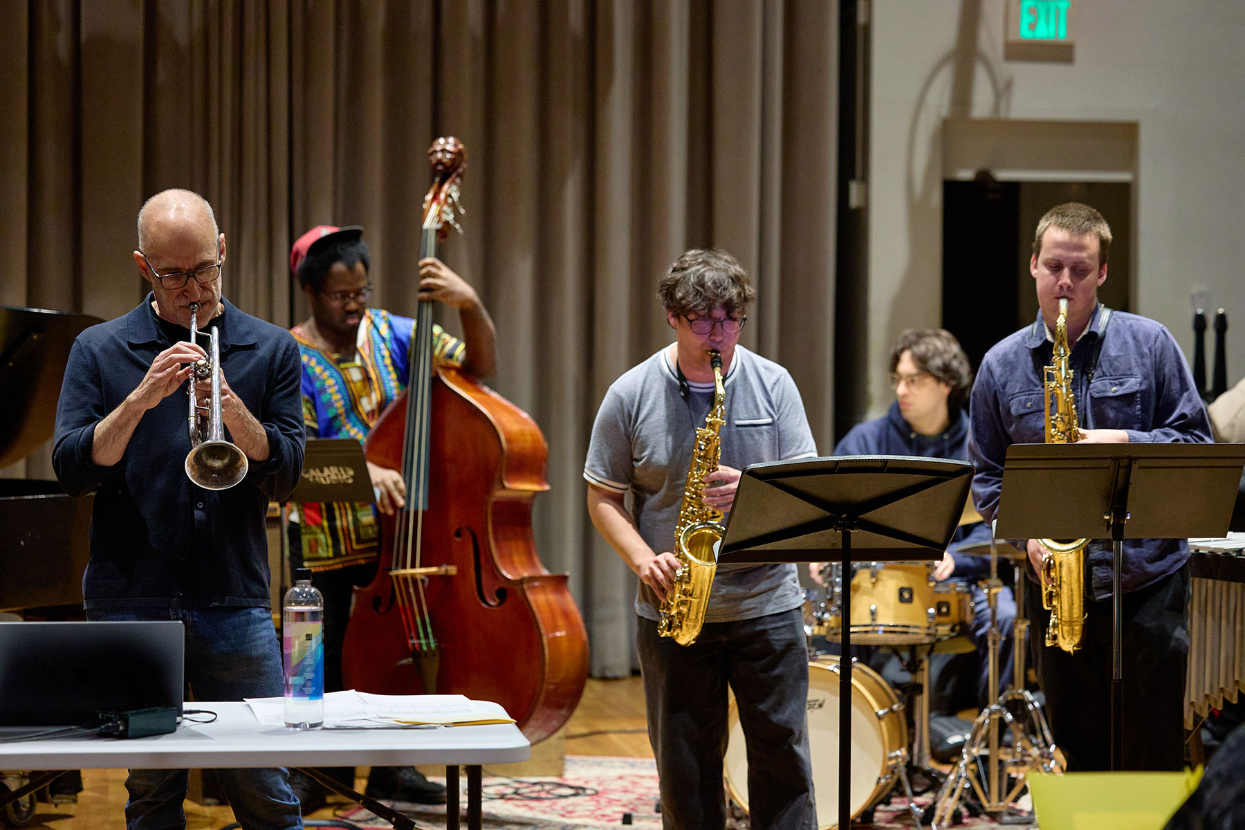 A jazz ensemble featuring a trumpeter, two saxophonists, an upright bass player, and a drummer rehearsing indoors against a tan curtain backdrop.