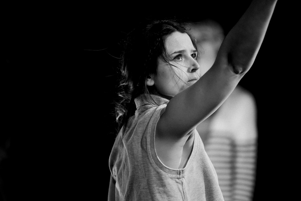 A black-and-white photo of a performer raising one arm while looking upward during a stage performance. Another figure appears out of focus in the background.