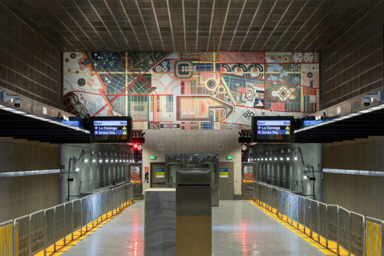A view down the platform of a Los Angeles Metro D Line station featuring a large colorful geometric mural spanning the far wall above the tracks, with digital train signs and symmetrical station architecture in the foreground.