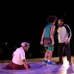 Three performers on a stage, with one kneeling and two standing, engaged in an intense moment.
