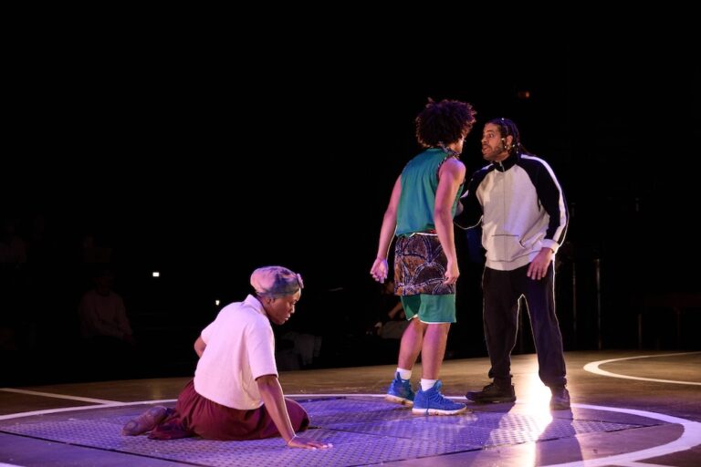 Three performers on a stage, with one kneeling and two standing, engaged in an intense moment.