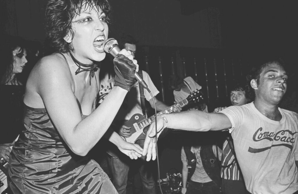 Black-and-white photograph of punk band The Bags performing live in Los Angeles in 1978. Lead singer Alice Bag sings intensely into a microphone while audience members and bandmates crowd closely around the stage during an energetic performance.