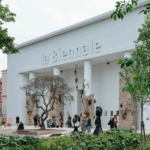 Building with "la Biennale" in raised letters, featuring tall white columns, a tree sculpture, and people in front.