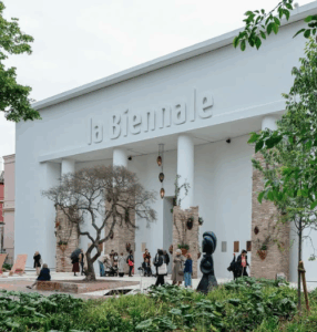 Building with "la Biennale" in raised letters, featuring tall white columns, a tree sculpture, and people in front.
