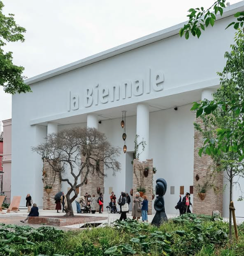 Building with "la Biennale" in raised letters, featuring tall white columns, a tree sculpture, and people in front.