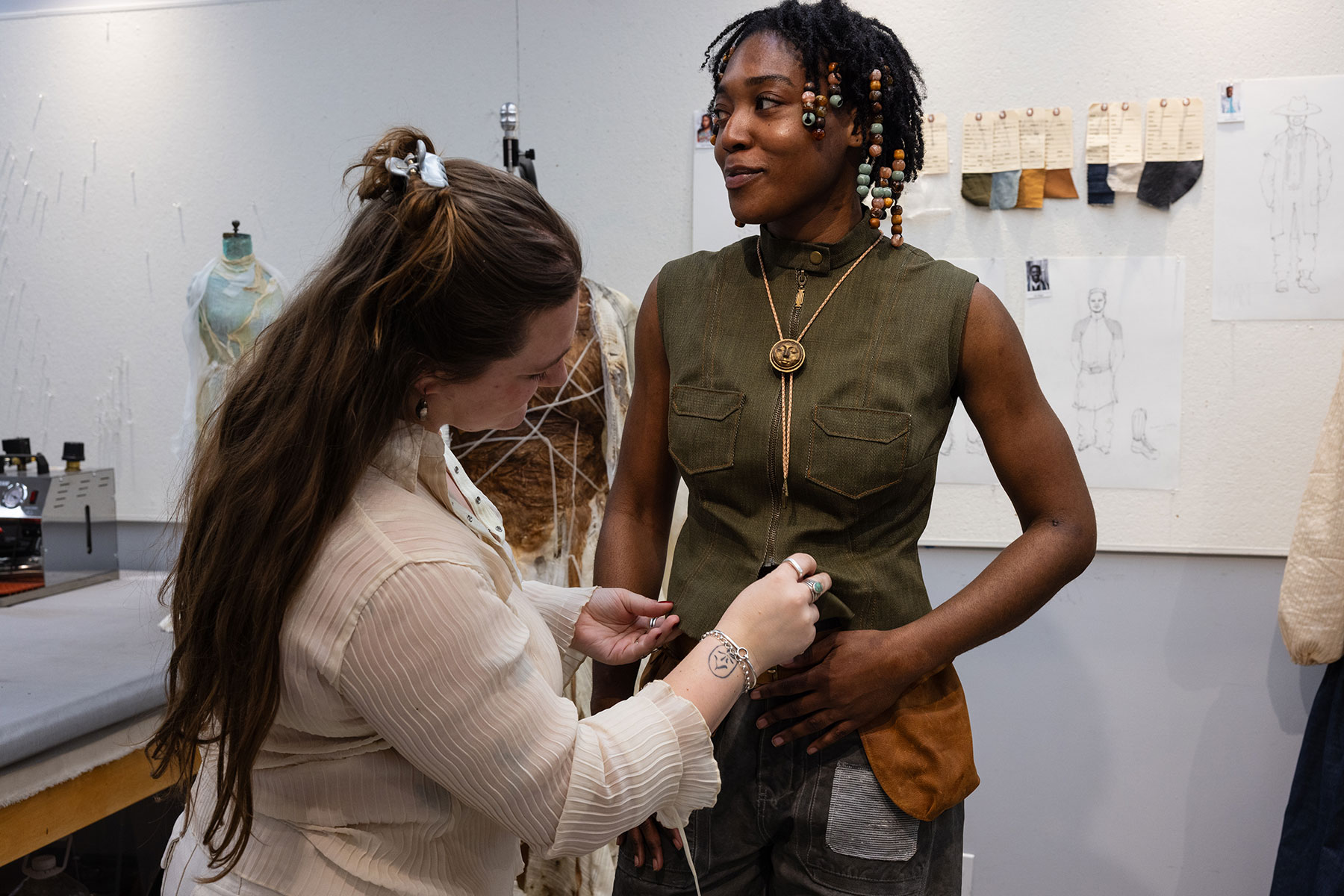 A fashion designer measures a smiling model wearing a green sleeveless shirt and a bolo tie in a studio setting with sketches on the wall.