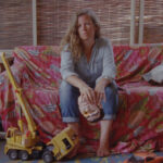 Image of a woman in denim sitting on a couch covered with a red-patterned cover. A small bulldozer lays at her feet.